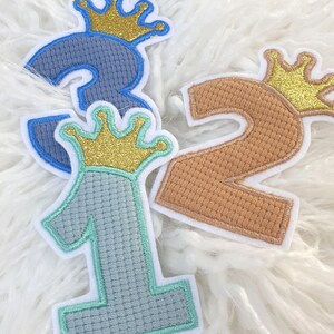 Könnte beinhalten: Gestickte Stoffaufnäher mit den Zahlen 1, 2 und 3, jeweils mit einer goldenen Glitzerkrone versehen. Die Zahlen sind in Blau- und Beigetönen gehalten, mit einem hellblauen und mintgrünen Rand. Ideal zum Personalisieren von Kleidung oder Accessoires.