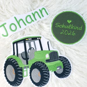 Könnte beinhalten: Gestickte Aufnäher mit einem grünen Traktor, dem Namen "Johann" in grüner Schrift und einem grünen Kreis mit "Schulkind 2026" und einem Herz. Der Traktor hat schwarze Reifen und einen grauen Innenraum. Alle Aufnäher sind auf weißem, flauschigem Hintergrund.