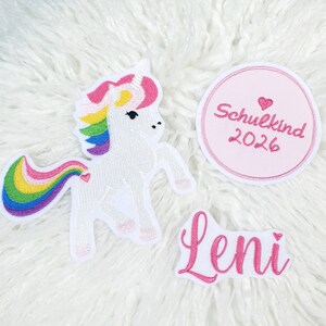 Könnte beinhalten: Gestickte Aufnäher mit einem weißen Einhorn mit Regenbogenmähne und -schwanz, einem rosa Kreis mit dem Text "Schulkind 2026" und einem rosa Namensaufnäher mit "Leni". Die Aufnäher befinden sich auf einer weißen, flauschigen Oberfläche.
