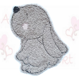 Könnte beinhalten: Graue, plüschige Hasenapplikation mit schwarzen Nahtdetails. Der Hase sitzt mit leicht geneigtem Kopf. Die Applikation hat ein strukturiertes Aussehen und einen hellrosa Akzent auf der Wange.