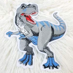 Könnte beinhalten: Gestickter Aufnäher mit einem grauen Tyrannosaurus Rex Dinosaurier mit blauen Akzenten. Der Dinosaurier hat ein rotes Maul und blaue Krallen. Der Aufnäher befindet sich auf weißem Hintergrund.