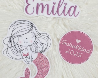 Puede incluir: Tres parches bordados: un parche con el nombre blanco y rosa que dice "Emilia", una sirena con una cola de purpurina rosa y un parche circular rosa con "Schulkind 2025".
