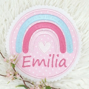 Könnte beinhalten: Ein runder, rosa Aufnäher mit einem Regenbogen-Design in Rosa- und Blautönen. Der Aufnäher zeigt den Namen "Emilia" in rosa Schrift und ein weißes Herz. Der Hintergrund ist rosa mit weißen Punkten und einem gewellten Rand.