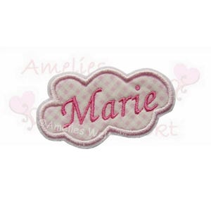 Könnte beinhalten: Ein rosa, wolkenförmiger, gestickter Aufnäher mit dem Namen "Marie" in Schreibschrift. Der Aufnäher hat ein hellrosa Vichy-Muster und eine dunkelrosa Umrandung. Der Text "Amelies W" ist ebenfalls sichtbar.