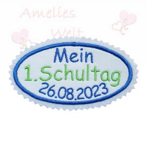 Könnte beinhalten: Ein bestickter ovaler Aufnäher mit blauem Rand und weißer Mitte. Der Text "Mein 1. Schultag 26.08.2023" ist in Blau und Grün gestickt. Der Aufnäher hat einen gewellten Rand.