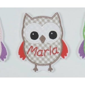 Könnte beinhalten: Ein aufgenähter Aufnäher in Eulenform mit einem karierten Muster in Beige und Weiß, roten Flügeln und dem Namen "Marla" in Rot gestickt. Die Eule hat große schwarze Augen und einen braunen Schnabel. Der Aufnäher ist wahrscheinlich für Kleidung oder Accessoires.