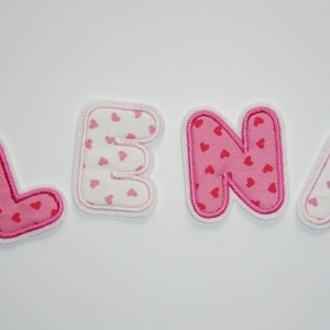 Könnte beinhalten: Vier Stoffbuchstaben buchstabieren den Namen "LENI". Die Buchstaben sind rosa mit einem Muster aus kleinen roten Herzen. Jeder Buchstabe hat einen weißen Rand und einen weißen Hintergrund. Die Buchstaben sind für Bastel- oder Personalisierungsprojekte konzipiert.