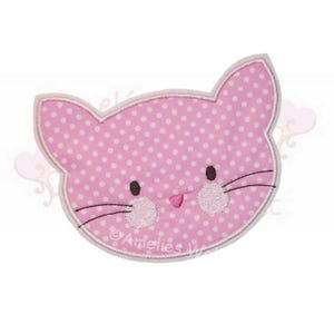 Könnte beinhalten: Ein rosa Applikation in Form eines Katzenkopfes mit weißen Punkten, schwarzen Augen und Schnurrhaaren. Die Katze hat eine rosa Nase und gestickte Wangen. Die Applikation ist weiß umrandet und trägt den Text "Amelies W".