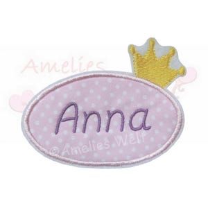 Könnte beinhalten: Ein rosa, ovales Patch mit weißen Punkten und dem Namen "Anna" in Lila gestickt. Eine goldene Krone mit weißen Akzenten sitzt über dem Oval. Der Patch hat einen weißen Rand und der Text "Amelies Welt" ist sichtbar.