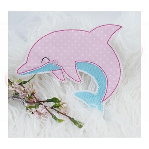 Könnte beinhalten: Ein rosa Delfin-Applikation mit weißen Punkten und einem hellblauen Bauch. Der Delfin hat ein lächelndes Gesicht und ist mit einem dunkelrosa Stich umrandet. Ideal für Bastel- und Nähprojekte.