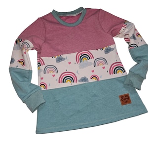 Könnte beinhalten: Ein langärmliges Oberteil mit Colorblock-Design. Das Oberteil hat einen rosa oberen Bereich, einen weißen mittleren Bereich mit Regenbogen- und Wolkenmustern und einen türkisfarbenen unteren Bereich. Die Ärmel sind türkis mit Regenbogenmustern.