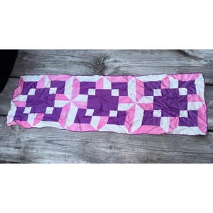 May include: A rectangular fabric runner featuring a repeating geometric quilt pattern in shades of purple, light pink, and white. The design consists of three central purple squares surrounded by alternating pink and white triangular and square shapes.