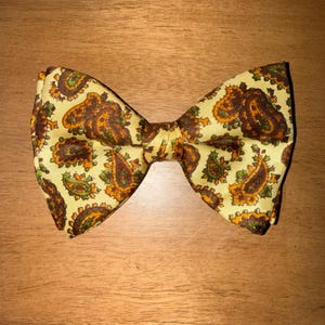 May include: A pre-tied bow tie featuring a light yellow background with a repeating paisley pattern in shades of dark brown, orange, and muted green. The fabric has a subtle sheen and a structured shape with pointed ends on both sides.