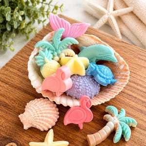 May include: A collection of small, pastel-colored soaps shaped like tropical and marine items, including a pink mermaid tail, a turquoise palm tree, a yellow pineapple, a blue dolphin, a blue turtle, a purple sea urchin, and pink flamingos.