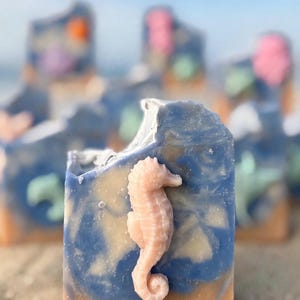 May include: A rectangular bar of soap featuring a marbled blue and white top section and a solid tan base. A three-dimensional, light pink seahorse figure is embedded on the front surface of the soap, which has a textured, uneven top edge.
