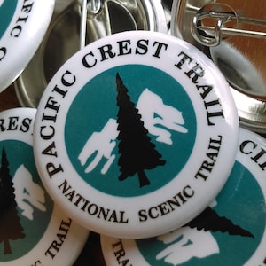 May include: A collection of white round buttons with a blue and green design. The buttons feature the text "Pacific Crest Trail" and a stylized image of a pine tree and mountains.