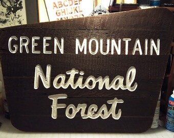 May include: A wooden sign with white lettering that reads "GREEN MOUNTAIN NATIONAL FOREST". The sign is dark brown and has a rustic look.