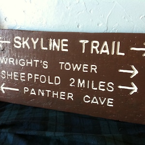 May include: A brown wooden sign with white painted text indicating directions to different trails. The sign reads "Skyline Trail", "Wright's Tower", "Sheepfold 2 miles", and "Panther Cave".