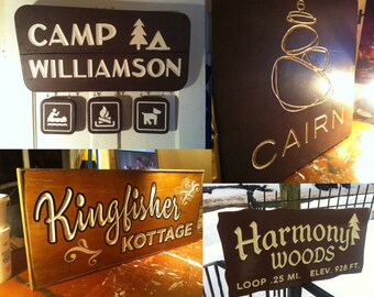 May include: Four wooden signs with different text and designs. The first sign says "Camp Williamson" with a tree and triangle symbol. The second sign says "Cairn" with a stylized image of a cairn. The third sign says "Kingfisher Kotttage" with a decorative flourish. The fourth sign says "Harmony Woods" with a tree symbol and the text "Loop .25 Mi. Elev. 928 Ft."