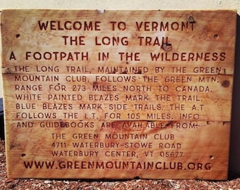 May include: A wooden sign with the text "Welcome to Vermont The Long Trail A Footpath in the Wilderness" and information about the Green Mountain Club, the Long Trail, and its location. The sign is brown with red text.