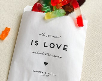 May include: White paper bag with the text "all you need is LOVE and a little candy" and a heart symbol. Below the text is "SANDRA & SIMON 10.8.22". The bag is filled with red, green, and orange gummy worms.
