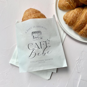 May include: A white paper treat bag featuring black text that reads A BABY IS BREWING, CAFÉ Bébé, and STELLA'S BABY SHOWER 09.21.24, with a line drawing of a cafe storefront. A golden-brown croissant rests inside the bag, with more on a white plate.