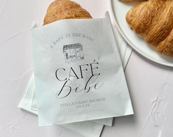 May include: A white paper treat bag featuring black text that reads A BABY IS BREWING, CAFÉ Bébé, and STELLA'S BABY SHOWER 09.21.24, with a line drawing of a cafe storefront. A golden-brown croissant rests inside the bag, with more on a white plate.