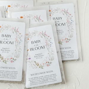 May include: A set of white envelopes with floral designs and the text "Baby in Bloom" and "Angelica's Baby Shower 07.21.25". Each envelope contains wildflower seeds and instructions for planting.