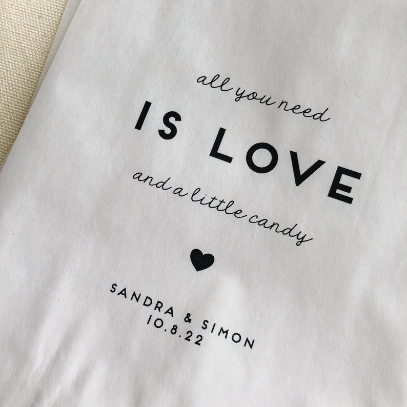May include: White paper bag with black text that reads "all you need is LOVE and a little candy". A small black heart is below the text. The text "SANDRA & SIMON 10.8.22" is below the heart.