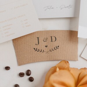 May include: A brown paper coffee cup sleeve with the initials "J&D" and a small heart design. The initials are surrounded by a simple leafy design.