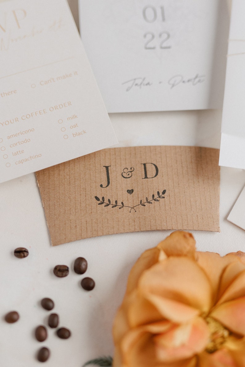 May include: A brown paper coffee cup sleeve with the initials "J&D" and a small heart design. The initials are surrounded by a simple leafy design.