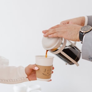 May include: A person pours coffee from a French press into a white paper cup with a brown paper sleeve that says "J & D" with a heart and a floral design.