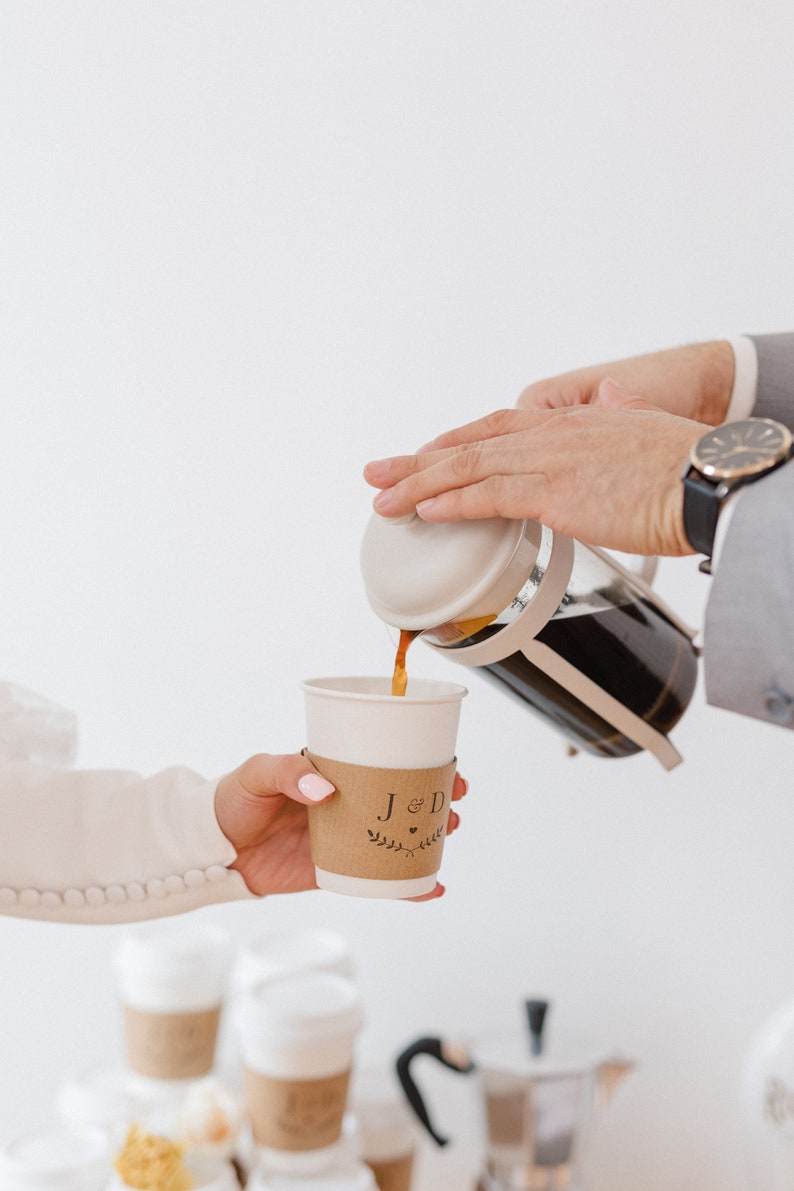 May include: A person pours coffee from a French press into a white paper cup with a brown paper sleeve that says "J & D" with a heart and a floral design.