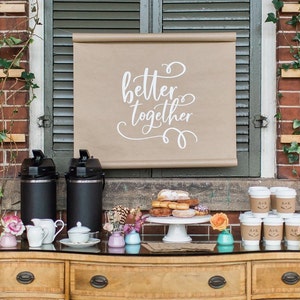 May include: A coffee and donut bar set up outdoors with a sign that reads "better together". There are two black coffee carafes, a white cake stand with donuts, and several small cups with personalized labels.