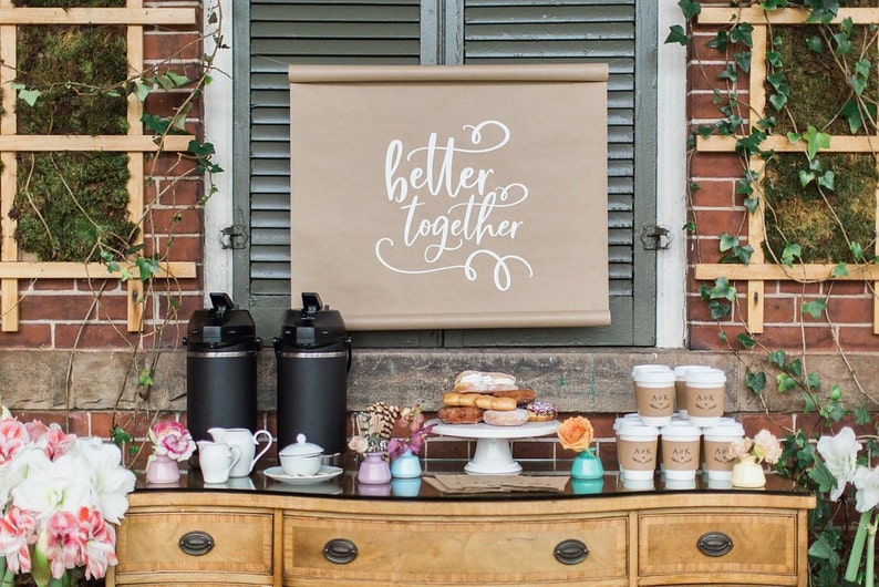 May include: A coffee and donut bar set up outdoors with a sign that reads "better together". There are two black coffee carafes, a white cake stand with donuts, and several small cups with personalized labels.