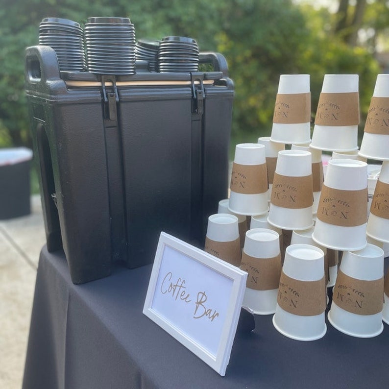 May include: A black cooler filled with disposable coffee cups and lids. The cooler is on a black table cloth with a white sign that says "Coffee Bar".