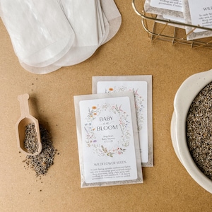 May include: A white envelope with a floral design and the text "Baby in Bloom" and "Wildflower Seeds". The envelope is filled with small brown seeds. The text also includes the date "01.21.23" and the words "America's Baby Shower".