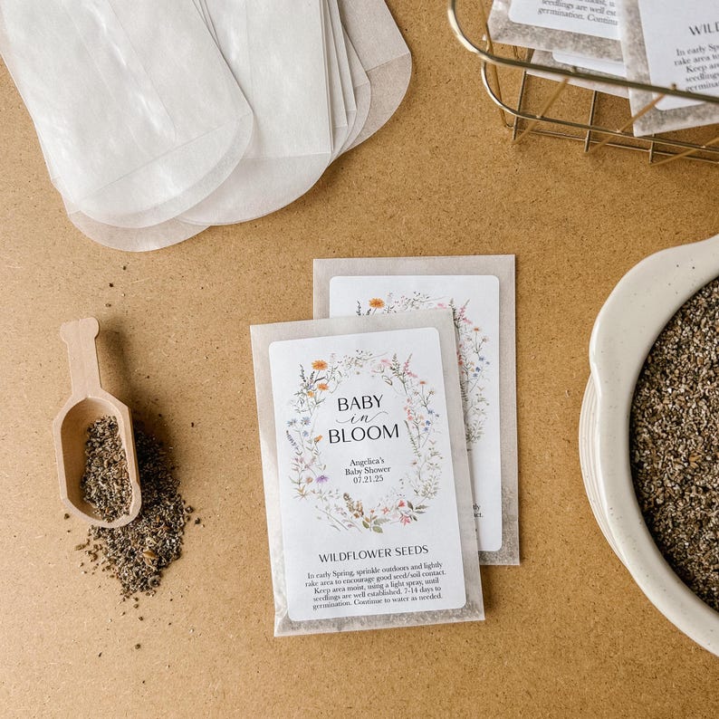 May include: A white envelope with a floral design and the text "Baby in Bloom" and "Wildflower Seeds". The envelope is filled with small brown seeds. The text also includes the date "01.21.23" and the words "America's Baby Shower".