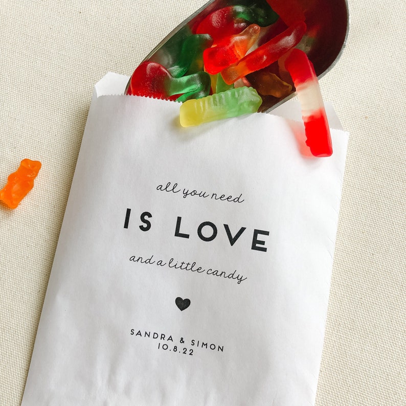 May include: White paper bag with the text "all you need is LOVE and a little candy" and a heart symbol. Below the text is "SANDRA & SIMON 10.8.22". The bag is filled with red, green, and orange gummy worms.