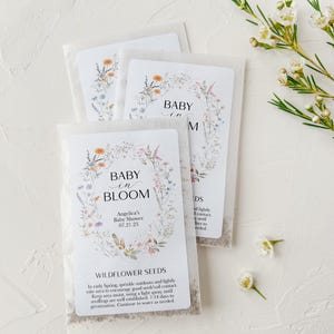 May include: White envelopes with wildflower seeds inside. The envelopes have a floral design and the text "Baby in Bloom" with a date and name. The text "Wildflower Seeds" is also included with instructions on how to plant the seeds.