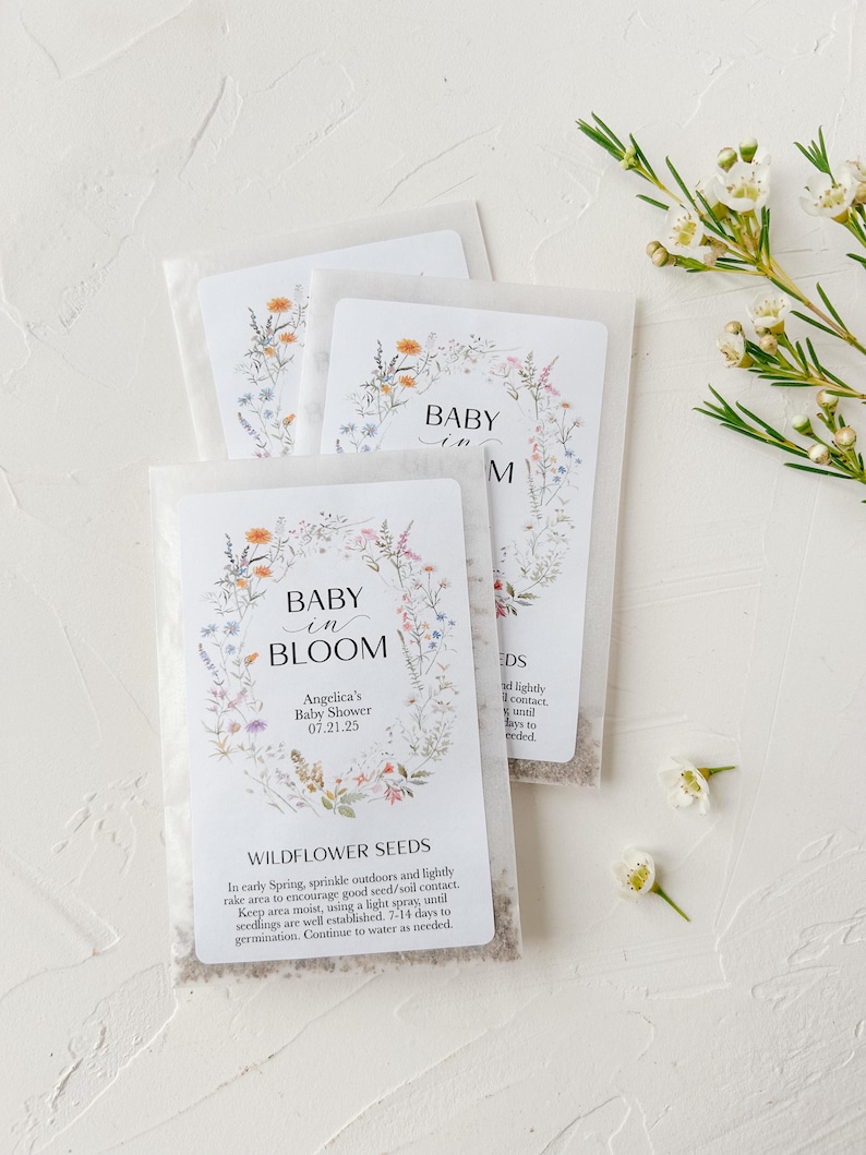May include: White envelopes with wildflower seeds inside. The envelopes have a floral design and the text "Baby in Bloom" with a date and name. The text "Wildflower Seeds" is also included with instructions on how to plant the seeds.