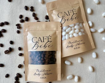 May include: Two kraft paper pouches with clear windows, each labeled CAFÉ Bébé and Martina's Baby Shower 10.15.26. One pouch contains dark roasted coffee beans, and the other contains white jelly beans, surrounded by scattered coffee and jelly beans.