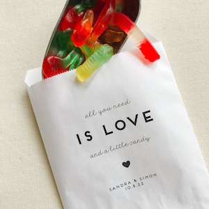 May include: White paper bag with the text "all you need is LOVE and a little candy" and a small heart. Below the text is "SANDRA & SIMON 10.8.22". The bag is filled with red, green, and yellow gummy worms.