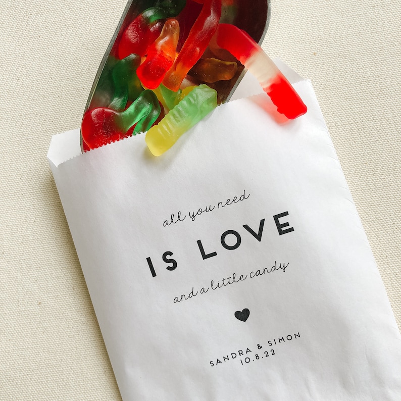 May include: White paper bag with the text "all you need is LOVE and a little candy" and a small heart. Below the text is "SANDRA & SIMON 10.8.22". The bag is filled with red, green, and yellow gummy worms.