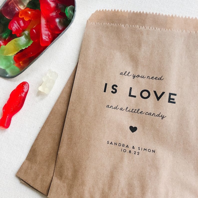 May include: Brown paper favor bag with the text "all you need is LOVE and a little candy" printed in black. A small black heart is below the text. The text "SANDRA & SIMON 10.8.22" is printed below the heart.