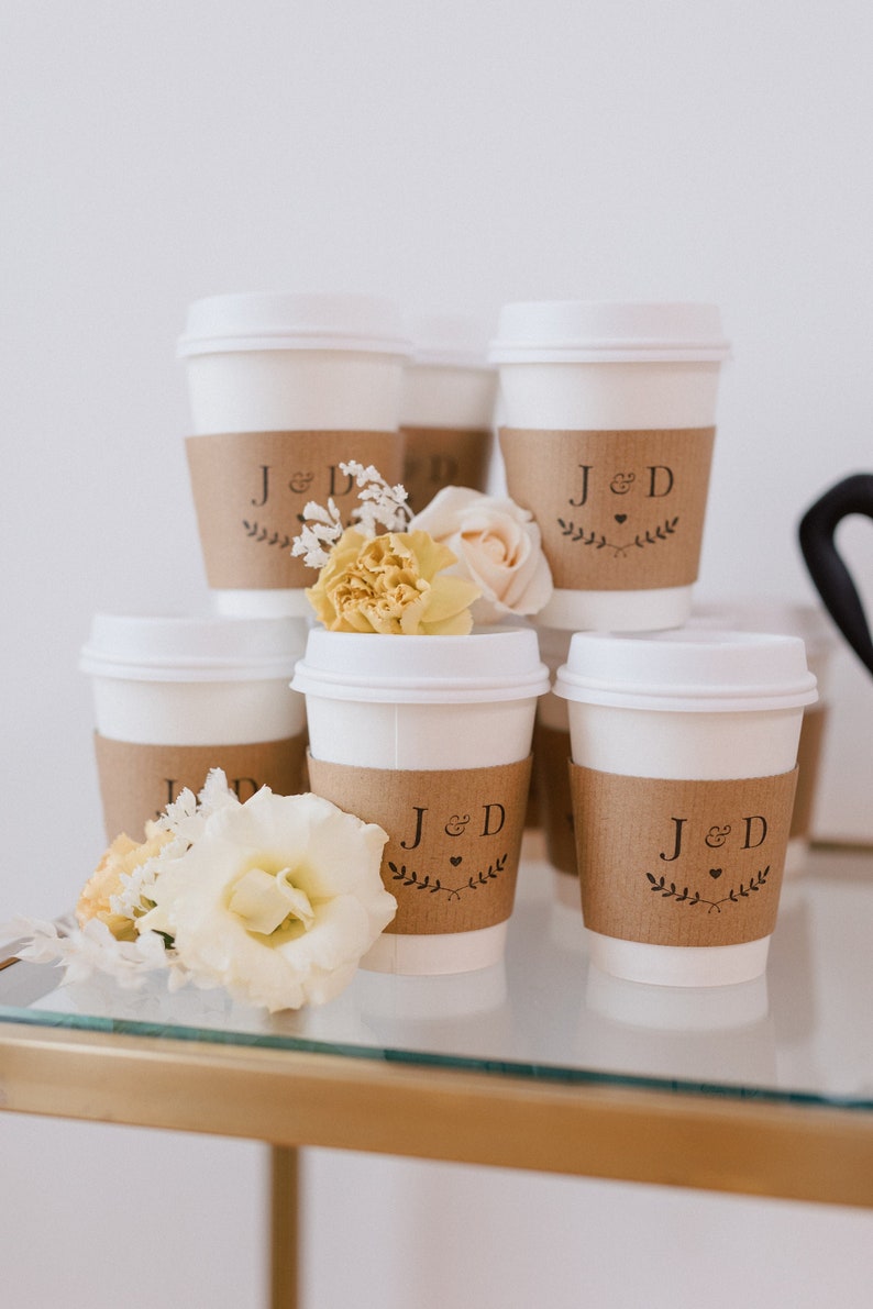May include: White coffee cups with brown paper sleeves decorated with the initials "J & D" and a small heart. The sleeves are adorned with a simple floral design. A bouquet of yellow and white flowers sits in front of the cups.