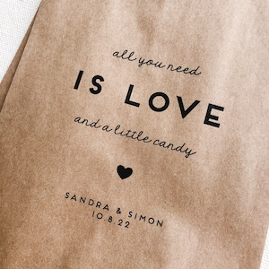 May include: A brown paper bag with the text "all you need is LOVE and a little candy" printed on it. A small heart is below the text. The names "SANDRA & SIMON" and the date "10.8.22" are printed below the heart.