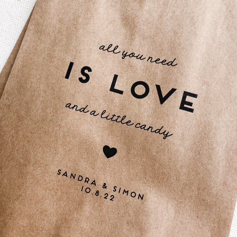 May include: A brown paper bag with the text "all you need is LOVE and a little candy" printed on it. A small heart is below the text. The names "SANDRA & SIMON" and the date "10.8.22" are printed below the heart.