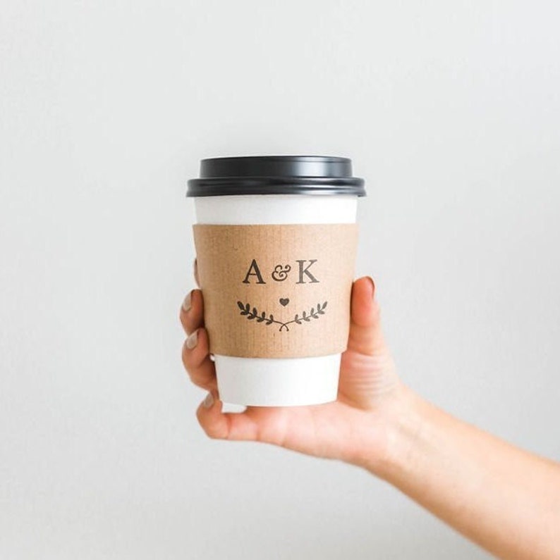 May include: A white to-go coffee cup with a brown paper sleeve. The sleeve has the initials "A & K" with a small heart and a leafy design.