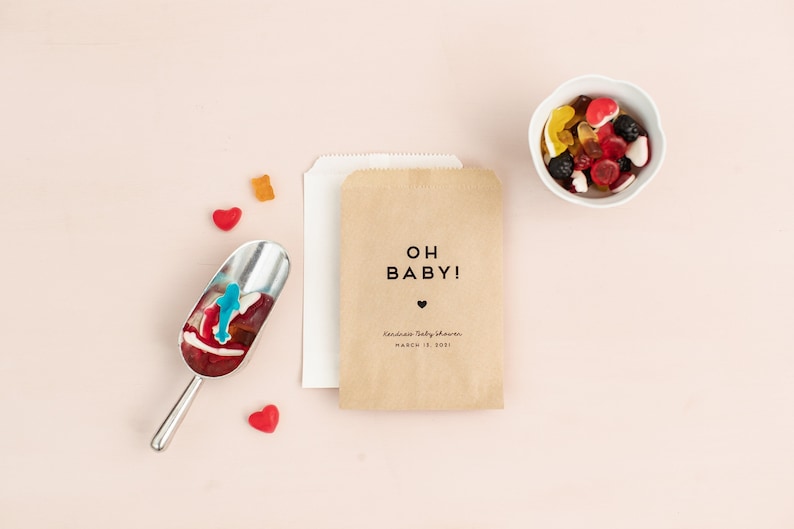 Puede incluir: Una bolsa de papel marr&oacute;n con el texto OH BABY! y un peque&ntilde;o coraz&oacute;n impreso en negro, acompa&ntilde;ada de una bolsa de papel blanca, una pala de metal con caramelos de goma variados y un cuenco blanco lleno de diversos dulces coloridos.